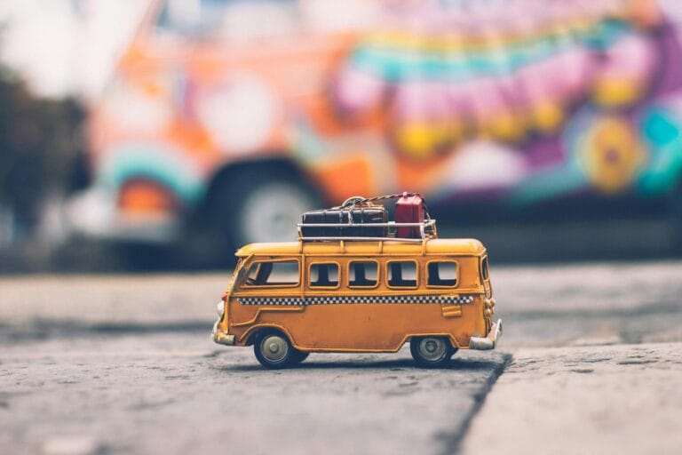 Toy yellow van with luggage on the roof parked on a street, with a colorful blurred vehicle in the background.